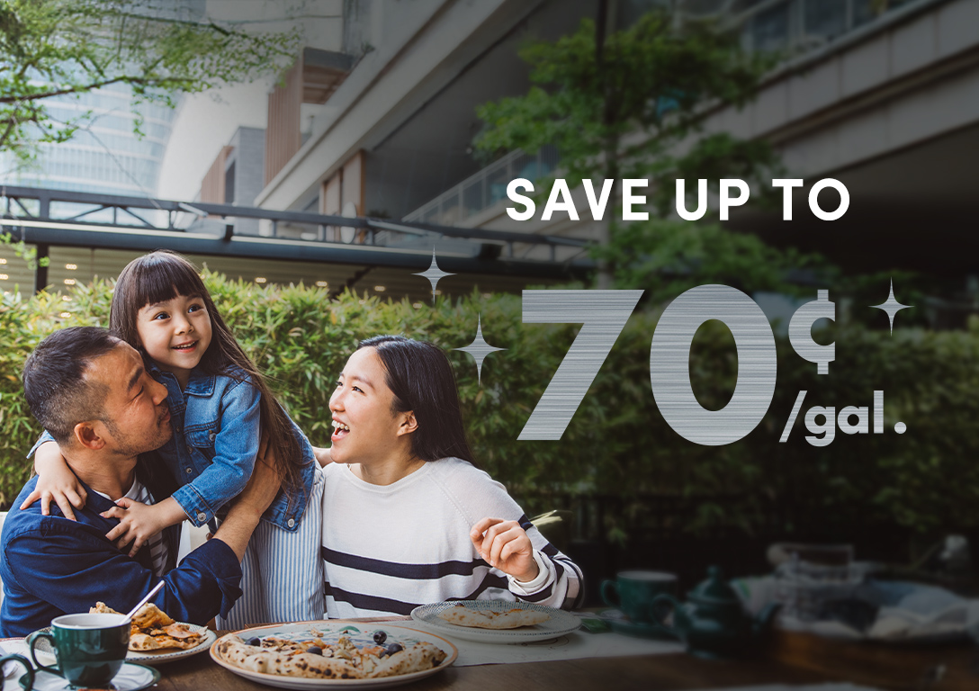A family enjoys a meal outdoors, surrounded by greenery, with dishes of food and drinks on the table. A promotional text reads 'Save up to 70¢/gal.'